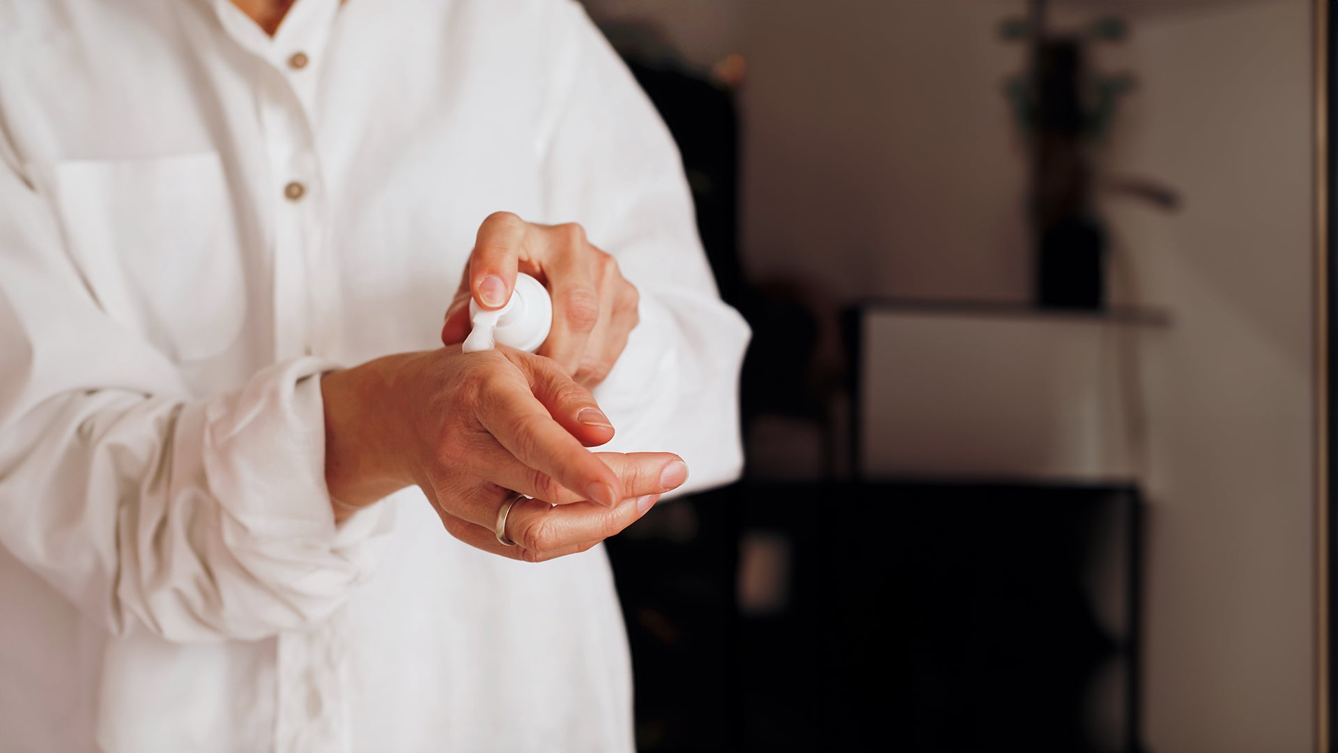 Woman in white shirt with buttons applying lotion product to right hand.