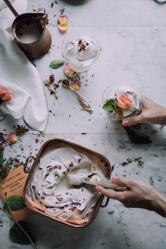 A batch of skin lotion being made by a person. Their two hands are shows from the right side of the frame scooping some product and filling a jar with other floral ingredients..